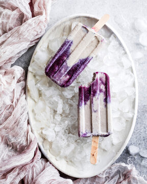 Two purple and cream popsicles on a bed of ice in a white dish, with a pink cloth nearby.