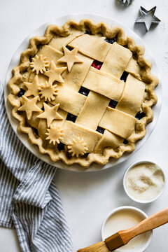 A decorative lattice-topped pie with star and sun-shaped cutouts, surrounded by kitchen utensils and ingredients on a white surface.
