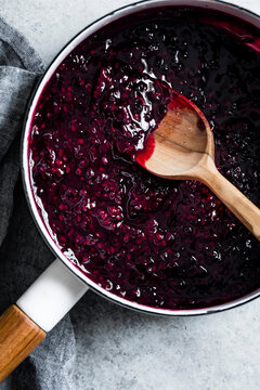 A saucepan filled with dark berry jam being stirred with a wooden spoon on a gray surface.