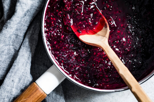 A saucepan filled with rich, dark berry jam and a wooden spoon on a gray cloth background.