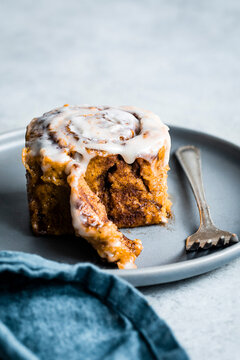 A cinnamon roll with icing on a gray plate, accompanied by a fork and a blue napkin.