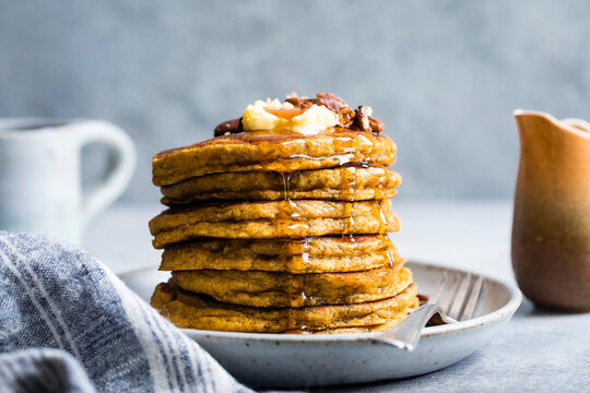A stack of pancakes topped with butter and nuts, drizzled with syrup, on a plate with a fork and linen napkin nearby.