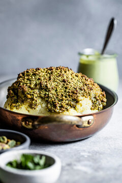 A roasted cauliflower head coated with a green crust, served in a copper pan with a creamy dip in the background.
