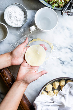 A person prepares dough circles on a marble countertop, surrounded by flour, a rolling pin, and filling ingredients.