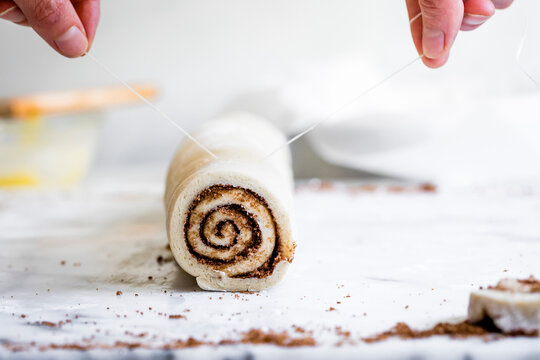 A person using dental floss to cut a rolled cinnamon dough on a white surface.