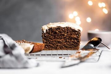 A sliced loaf of chocolate cake with white frosting on a cooling rack, surrounded by a knife and blurred lights in the background.