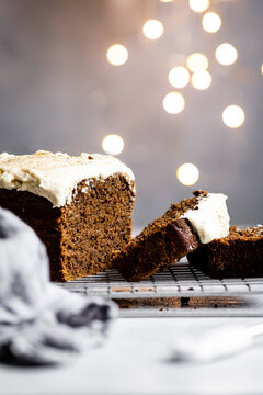 A sliced gingerbread cake with creamy white frosting on a cooling rack, set against a blurred background with glowing lights.