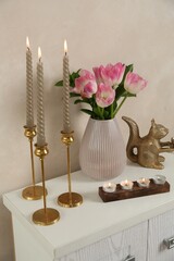 Elegant candlesticks with burning candles, tulips in vase and other decor on dresser near beige wall
