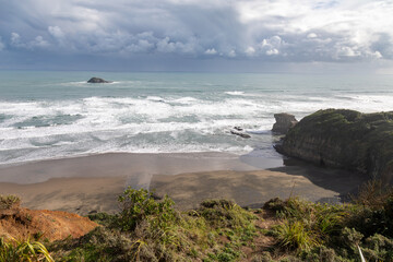 Powerful waves crash onto a sandy beach beside rugged cliffs under a dramatic cloudy sky on the coast of  Muriwai, Auckland, New Zealand