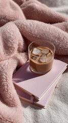 Aerial view of chilled coffee resting on pastel notebooks alongside soft blanket, embodying serene morning comfort