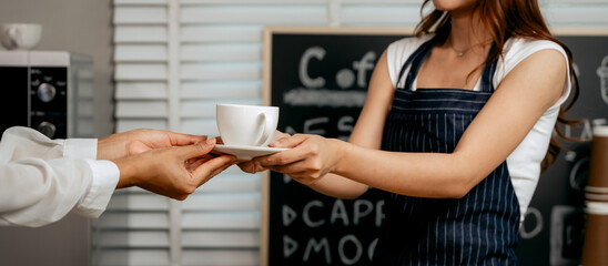 A young Asian female barista in a striped apron and orange shirt stands in a cozy modern café, smiling warmly as she hands a freshly made coffee cup to a woman customer.