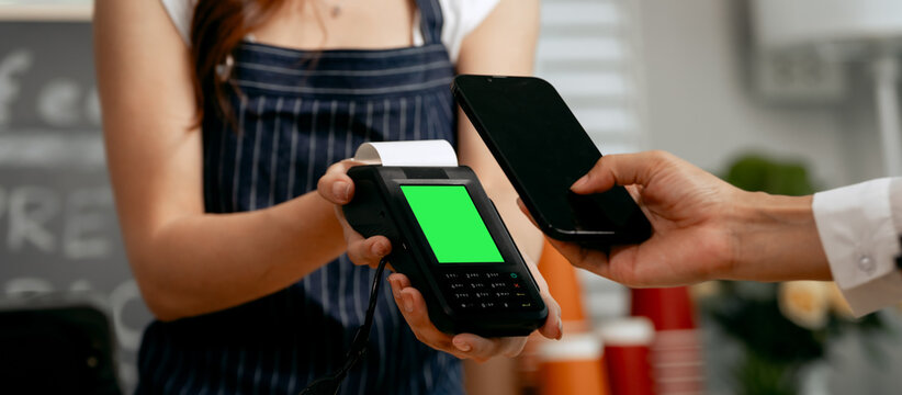 A young Asian female barista in a striped apron and orange shirt assists a woman customer making a quick, contactless payment via mobile QR code at the POS in a cozy modern cafe. - Powered by Adobe