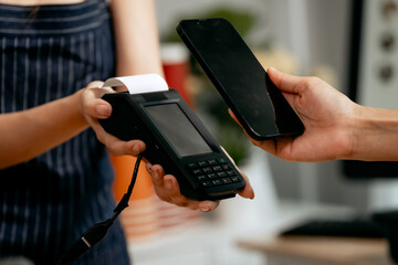 A young Asian female barista in a striped apron and orange shirt assists a woman customer making a quick, contactless payment via mobile QR code at the POS in a cozy modern cafe.