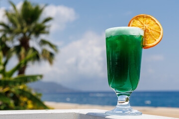 Vibrant green cocktail with an orange slice garnished on rim of a tall glass, set against tropical beach backdrop with palm trees and clear blue ocean. Copy space. Holiday travel.
