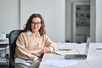Young happy pretty professional business woman at workplace. Smiling female company office worker, entrepreneur or businesswoman executive sitting at work desk, looking at camera, portrait.