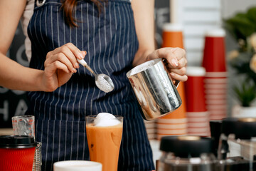 A young female barista in a striped apron prepared iced beverage Thai tea with whipped cream in cozy cafe, surrounded by coffee grinders, takeaway cups, pastries, and chalkboard menu in background.