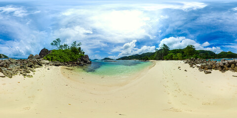 A serene beach with rocky formations and soft sand, leading into clear, calm waters with lush greenery in the background. Port Glaud Lagoon. Seychelles. VR 360.