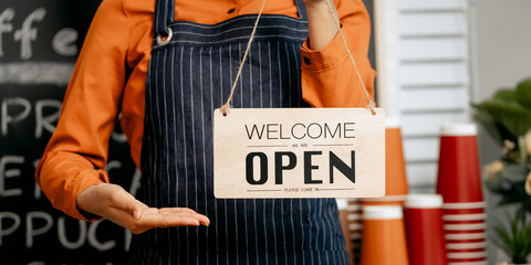 A smiling young Asian female barista in a striped apron and orange shirt holds a "Welcome Open" sign in a cozy modern café, surrounded by coffee equipment, takeaway cups, and pastries.