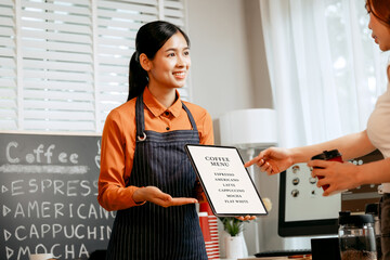 A young Asian female barista in modern cafe assists woman customer ordering latte, fresh coffee...