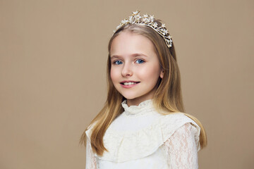 Young girl with a tiara, smiling brightly against a soft beige background, wearing a white lace dress, capturing a joyful, fairy tale essence with elegance and grace