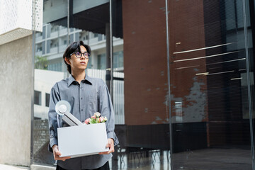 Young asian man in office shirt holding a box filled with personal belongings after losing his job,...