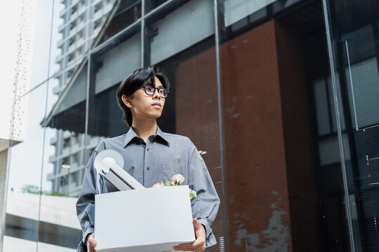 Young asian man in office shirt holding a box filled with personal belongings after losing his job, walking outside an office building with a serious and disappointed expression, unemployment.