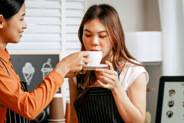 Two young Asian female baristas in striped aprons and orange shirts enjoy teaching each other how to make latte coffee, smell aroma, and taste coffee and croissants together in cozy modern café.