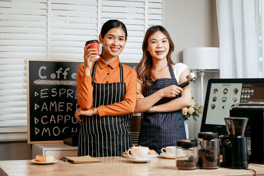 Two young Asian female baristas in striped aprons and orange shirts enjoy teaching each other how to make latte coffee, smell aroma, and taste coffee and croissants together in cozy modern café.
