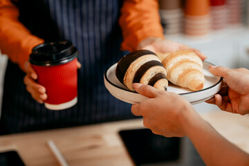 A young Asian female barista in modern cafe assists woman customer ordering latte, fresh coffee beans, dark roast, croissant, completing the purchase with mobile payment at the POS using QR code.
