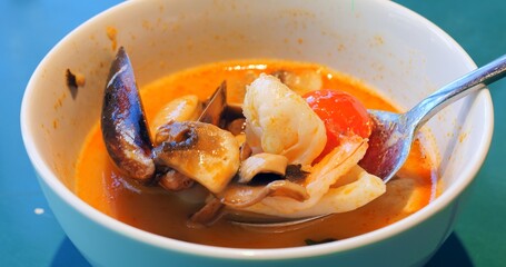 Spoon mixing and showing ingredients of a tom yum soup, a traditional spicy and sour thai soup, with seafood like shrimps, mussels, squid, and vegetables like mushrooms and tomatoes