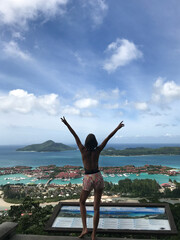 Young woman standing back to camera facing spectacular tropical view of island Mahe, Seychelles. Beautiful model with raised arms is enjoying traveling  on tropical destination.