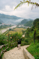 A person descends a stone path with arms raised, overlooking a misty valley, river, village, and...