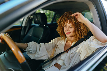 Smiling woman driving car with curly hair, wearing white clothes and seatbelt, enjoying sunny day...