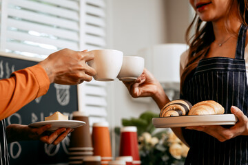 Two young Asian female baristas in striped aprons and orange shirts enjoy teaching each other how to make latte coffee, smell aroma, and taste coffee and croissants together in cozy modern café.