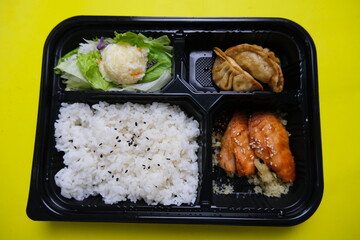 Japanese bento lunch box featuring steamed white rice with black sesame, crispy fried dumpling, glazed chicken, and fresh salad with potato topping.