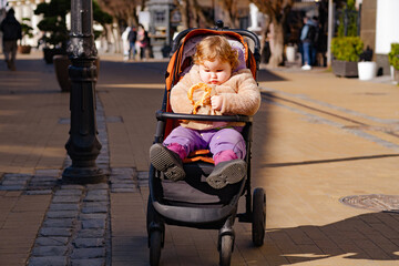 A child with curly hair happily eats a pretzel while sitting in a stroller in a bustling outdoor...