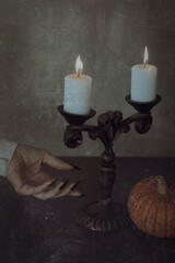 Female hand with an open palm and sharp nails reaches out to vintage candelabrum with burning candles and small pumpkin on the table, vertical image.