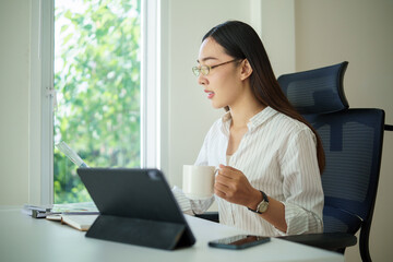 Young businesswoman holding a cup while analyzing reports on a tablet near a bright office window.