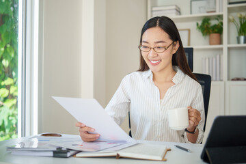 Young businesswoman holding a cup while analyzing reports on a tablet near a bright office window.