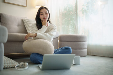 Obraz premium Young woman thoughtfully looking at laptop screen while sitting on the floor at home.
