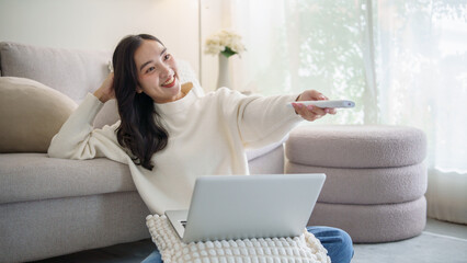 Cheerful Asian woman switching TV channels with a remote next to her laptop.