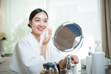 Smiling woman in bathrobe applying face cream during skincare routine.