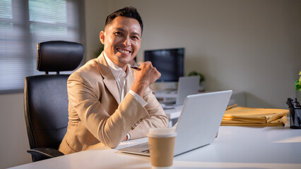 Confident businessman in beige suit working on laptop at office desk, smiling at camera with positive energy.