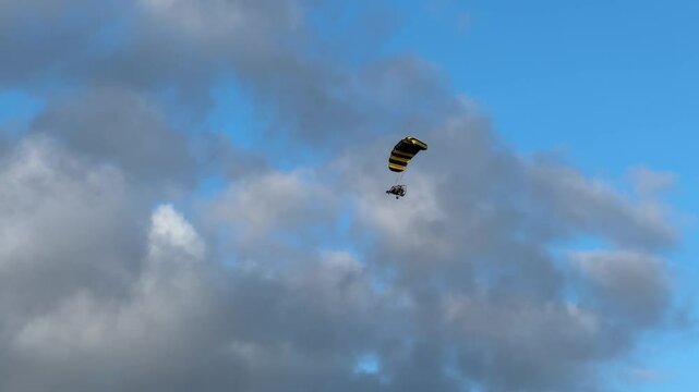 Ultralight powered parachute, with pilot and passenger, flies over head, but under clouds in late afternoon light.
