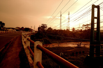 A scenic view of a bridge at sunset with power lines and a river below, surrounded by trees and a warm orange sky.