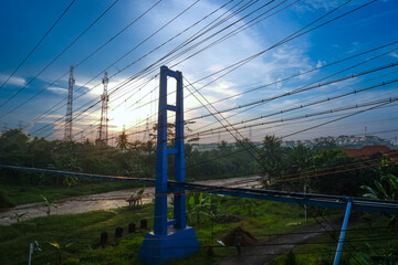 A blue tower structure with power lines against a sunrise sky, surrounded by greenery.
