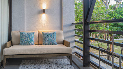 Outdoor seating area on the terrace. A sofa with soft cushions against the wall. The lamp is lit. Tiled floor.  A tropical park is visible behind the latticed railings. The resort hotel.