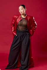Fashion asian woman in red leather jacket and black pants posing against a red background with...