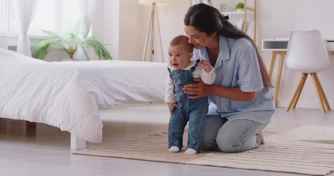 Portrait of happy cheerful mother sitting with her infant little cute child daughter supporting her in first steps at home. Toddler girl stepping on floor learning to walk with mom help.