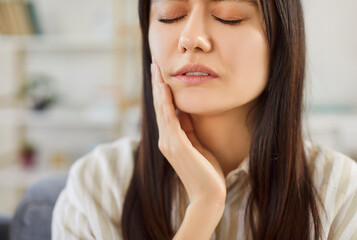 Closeup portrait of young unhappy upset woman sitting on the sofa in the living room at home suffering from toothache. Sad brunette girl in casual clothes holding her cheek in dental pain.